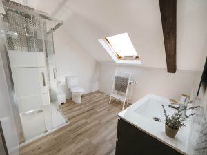 Interior del baño en la planta alta de Casa Bieiteiros con lavabo a la derecha, radiador toallero al fondo y ducha con mampara a la izquierda bajo techo abuhardillado