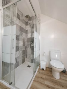 Vista interior del baño en la planta alta de Casa Bieiteiros, mostrando la taza del váter y la bañera con ducha bajo el techo abuhardillado de madera en Valdoviño